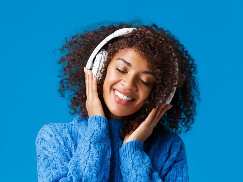 A curly-haired woman wearing a blue sweater, enjoying music on white headphones.